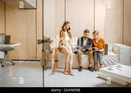Creative colleagues working on digital tablet and talking on phone on the couch indoors. Work in marketing agency or design studio concept Stock Photo