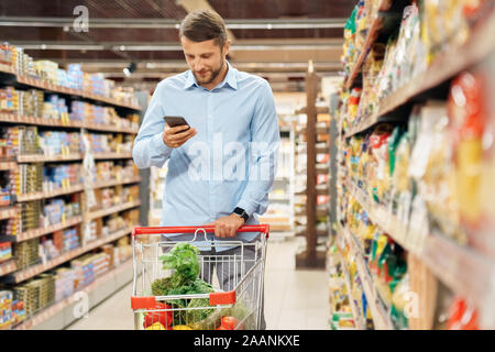 Young adult man shopping in supermarket and using smartphone Stock Photo