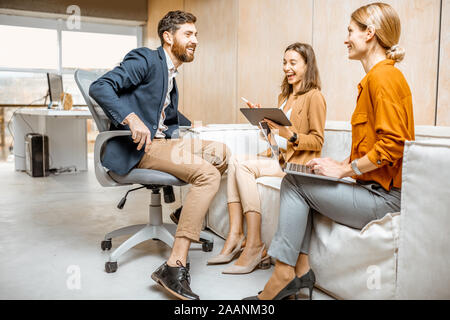 Playful man flirting with a young colleagues, women sitting and working on the couch in the office Stock Photo