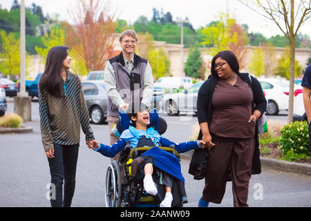 Disabled biracial boy in wheelchair walking along city street with father, siblings and caregiver, holding hands Stock Photo