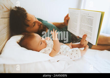 Family father and infant baby reading book fairytale laying on bed at home lifestyle dad and child daughter together parenthood childhood concept Fath Stock Photo