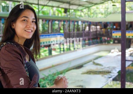 Bircaial teen girl or young woman standing on balcony overlooking crocodile show in Thailand Stock Photo