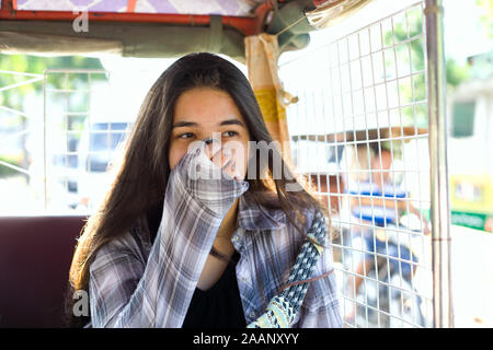 Biracial teen girl or young woman tourist covering nose and mouth from pollution while in tuk tuk taxi in Phnom Pehn, Cambodia Stock Photo