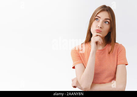 Thoughtful intrigued young fair-haired girl in striped t-shirt, touch chin and pouting curious, look upper left corner intrigued and unsure, making Stock Photo