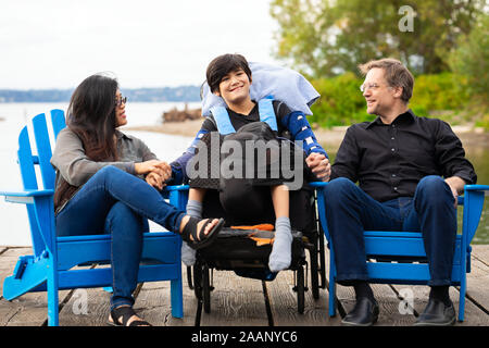 Multiracial couple sitting with disabled twelve year old son in wheelchair while sitting in blue adirondack chairs on wooden pier by lake on summer da Stock Photo