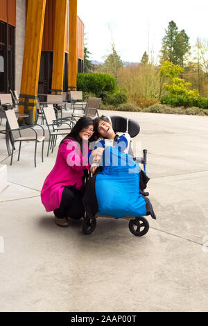 Asian mother in forties with eleven year old biracial disabled son in wheelchair outdoors Stock Photo