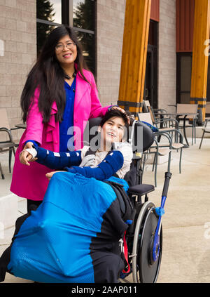 Asian mother in forties with eleven year old biracial disabled son in wheelchair outdoors Stock Photo