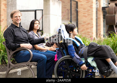 Multiracial family with special needs child sitting outdoors together on summer day. Child is sitting in wheelchair. Stock Photo