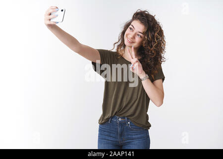 Cute tender young happy smiling armenian curly-haired girl 20s extend arm hold smartphone taking selfie tilt head show peace or victory gesture Stock Photo