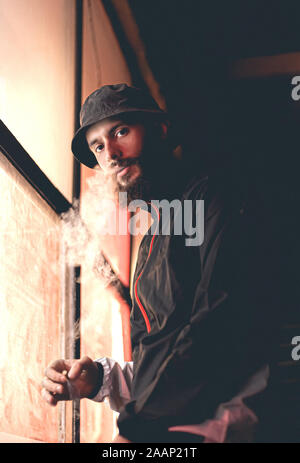 Bearded hipster smokes a cigarette at night urban style. Stock photo portrait of a smoking guy at night. Film effect. Stock Photo
