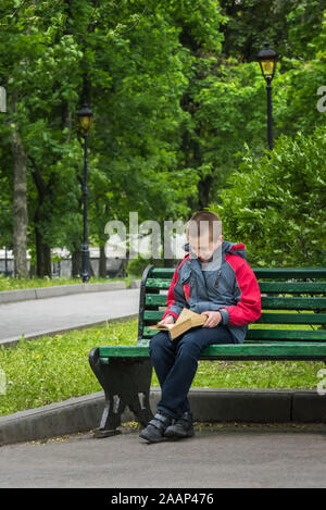 Smiling boy in casual clothing is reading a book. Boy is sitting alone on old wooden bench in park. Selective focus. Unfocused green park at backgroun Stock Photo