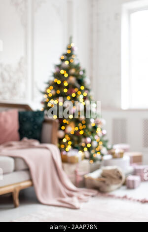 Blured photo of decorated wit garland and toys christmas tree standing next to white sofa. Stock Photo
