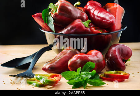 Red bulgarian pepper cut and intact with mint herb, slices and seeds all over the wooden table around big black glass bowl with harvest of vegetable w Stock Photo