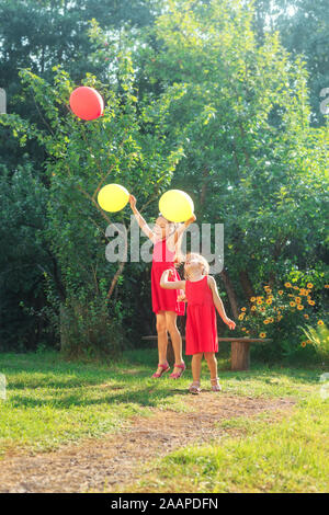 Two happy cute little sisters jumping with colorful toy balloons outdoors. Smiling kids having fun in green spring garden at warm summer day. Stock Photo