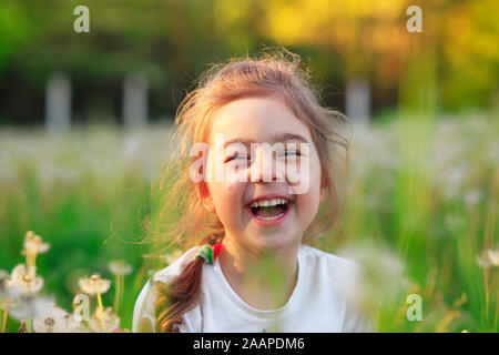 Beautiful little girl  laughing and playing with flowers in sunny spring park. Happy cute kid having fun outdoors at sunset. Stock Photo