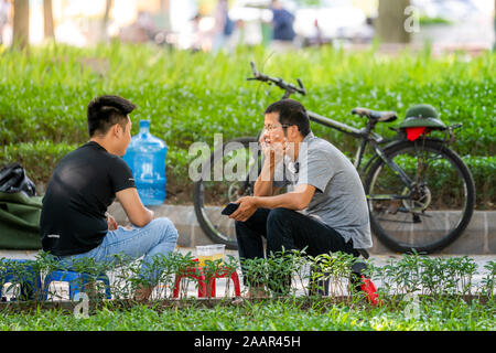 Hanoi, Vietnam - 12th October 2019: Two young asian men sit down in a park on plastic stools drinking juice and talking about their day Stock Photo