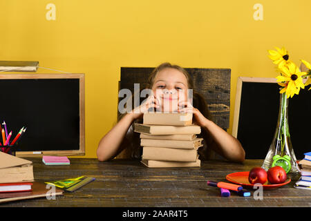 Schoolgirl with happy and funny face expression lies on pile of books. Kid and school supplies on yellow background. Childhood and back to school concept. Girl sits at desk with colorful stationery Stock Photo