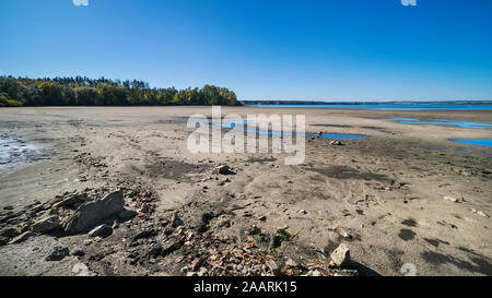 Dry landscape in extreme weather. Muddy bed of pond with some puddles in sand. Forest and water surface on horizon. Clear blue sky. Climate changes. Stock Photo