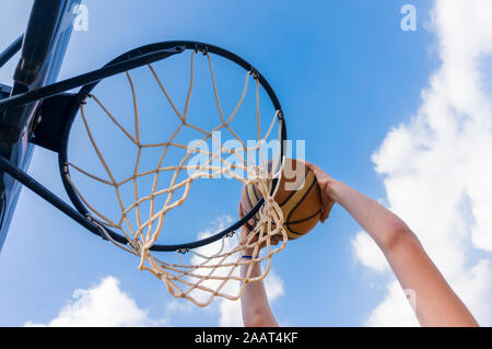 Young boy making a slam dunk in street basketball with blue sky and white clouds Stock Photo