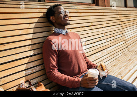 Young man sitting on wooden bench, eating hamburger, drinking coffee Stock Photo