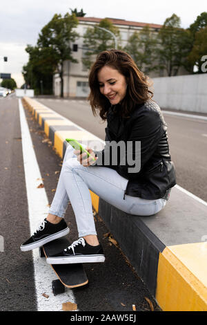 Smiling young woman with skateboard sitting on bollard using smartphone Stock Photo