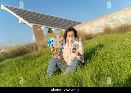 Portrait of smiling young woman sitting on a meadow with pinwheel looking at mobile phone Stock Photo