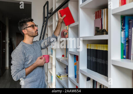 Portrait of young man with cup standing in front of bookshelves at home taking a book Stock Photo