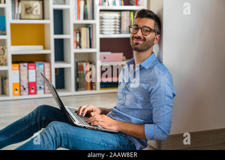 Portrait of smiling young man sitting on the floor at home using laptop Stock Photo