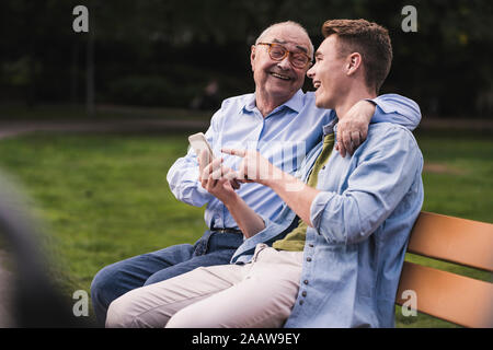 Senior man and grandson with smartphone sitting together on a park bench having fun Stock Photo