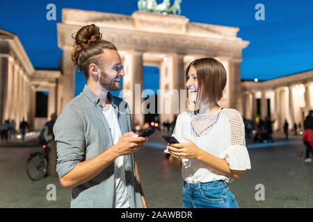 Happy couple using smartphones at Brandenburg gate at blue hour, Berlin, Germany Stock Photo