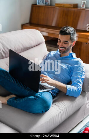 Smiling young man sitting on the couch at home using laptop Stock Photo