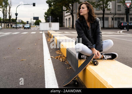 Young woman with skateboard sitting on bollard looking at distance Stock Photo