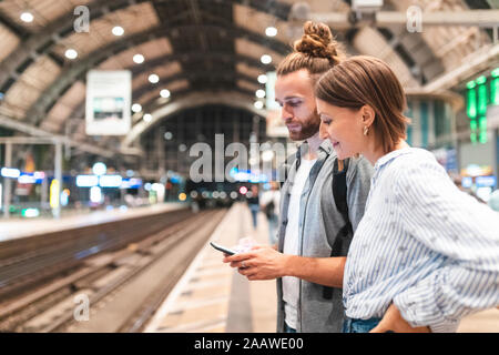 Young couple waiting for the train at the station and using smartphone, Berlin, Germany Stock Photo