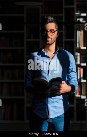 Portrait of young man with book standing in front of bookshelves at home Stock Photo