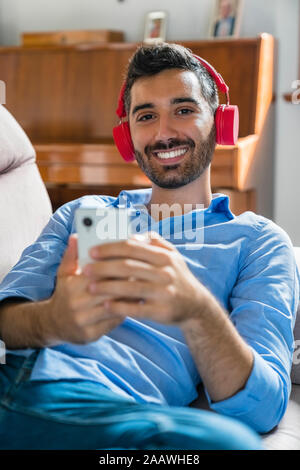 Portrait of smiling young man lying on the couch at home using smartphone and wireless headphones Stock Photo
