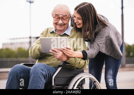 Senior man sitting in wheelchair and adult granddaughter looking together at digital tablet Stock Photo