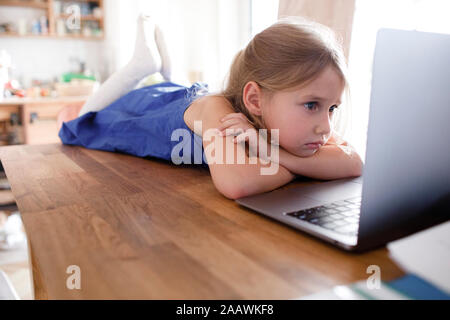 Portrait of sad little girl lying on  kitchen table at home looking at laptop Stock Photo