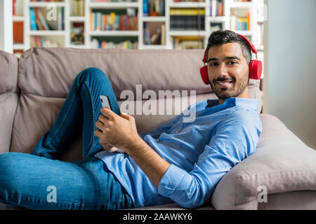 Portrait of smiling young man lying on the couch at home using smartphone and wireless headphones Stock Photo