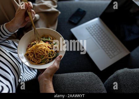 Mature woman with laptop eating homemade pasta dish on couch at home Stock Photo