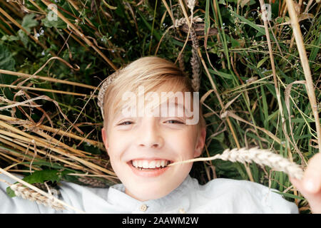 Portrait of smiling blond boy with oat ear in mouth lying in an oat field
