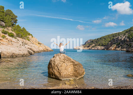 Redheaded woman with raised arms sitting on rock at the beach Stock Photo