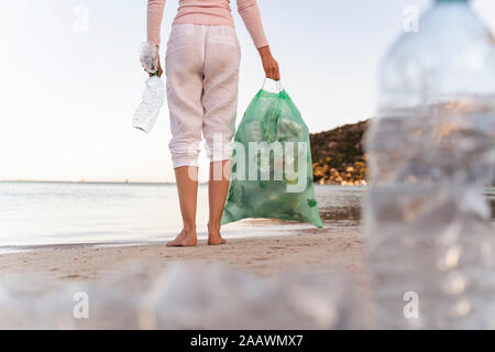 Back view of woman standing on the beach with bin bag of collected empty plastic bottles