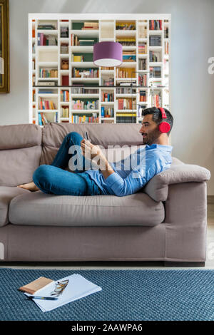 Smiling young man lying on the couch at home using smartphone and wireless headphones Stock Photo