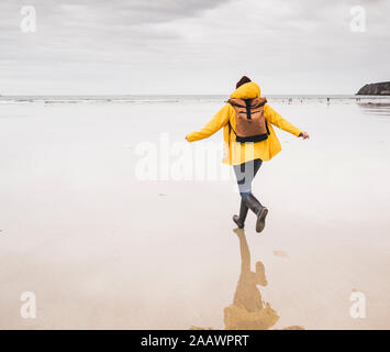 Young woman wearing yellow rain jacket and running at the beach, Bretagne, France Stock Photo