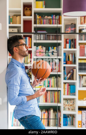 Pensive young man with basketball standing in front of bookshelves at home Stock Photo