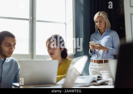 Colleagues discussing project, while mature woman is using smartphone in background Stock Photo