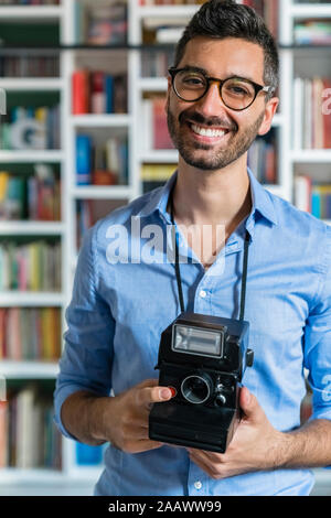 Portrait of happy young man with instant camera standing in front of bookshelves Stock Photo