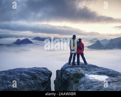 Lovely heterosexual Couple looking at far sunrise in heavy clouds. The dark night in foggy mountains ends. Stock Photo