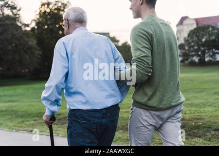 Back view of young man assisting his grandfather walking in a park Stock Photo