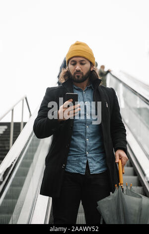 Portrait of man with umbrella standing on escalator looking at smartphone Stock Photo